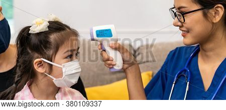 Panoramic Asian female doctor use infrared thermometer to take temperature to little girl child in living room while doctor visit at home. Home health care delivery and doctor visiting concept.