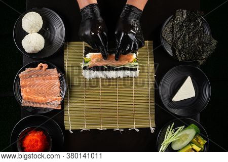 Process of making  California roll sushi by hands in black gloves on black background framed by plates with ingridients with salmon, cucumber, avocado, cream cheese, red masago caviar