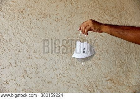 Male Hand Holding Up A White N95 Mask In Natural Light Against A Beige Textured Background