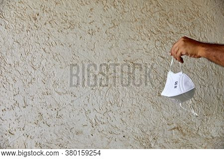 Hand Holding Up A White Kn95 Mask In Natural Light Against A Beige Textured Background With Lots Of 