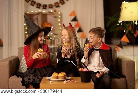 halloween, holiday and childhood concept - smiling boy and girls in costumes eating cupcakes and having fun at home at night