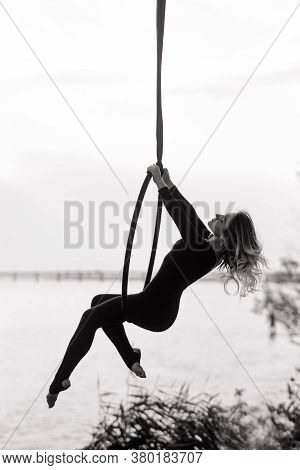 Woman Aerialist Performs Acrobatic Elements In Hanging Aerial Hoop Against Background Of River. Blac