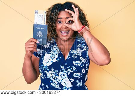 Young african american plus size woman holding united states passport and boarding pass smiling happy doing ok sign with hand on eye looking through fingers 