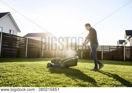 Spring Mowing The Lawn. Young Man In A Plaid Shirt And Jeans Mows Grass With A Lawn Mower In The Bac