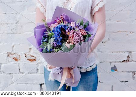 A Bouquet Of Flowers, Packed In Purple Paper In The Hands Of A Woman On A Light Background. Bouquet 