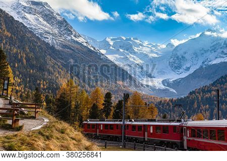 Stunning View Of Red Rhaetian Train Running Under The Morteratsch Glacier In Autumn With Blue Sky Cl