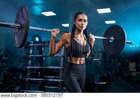 Side View Of Stunning Female Bodybuilder With Long Braids And Perfect Makeup Holding Barbell On Shou