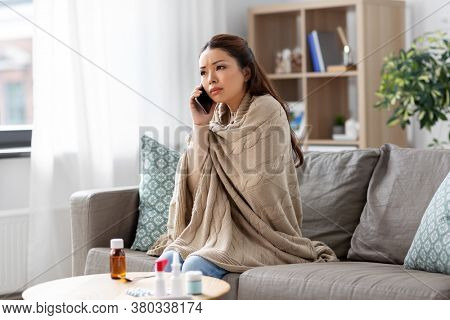 technology, health and cold concept - sick asian woman in blanket calling on smartphone at home