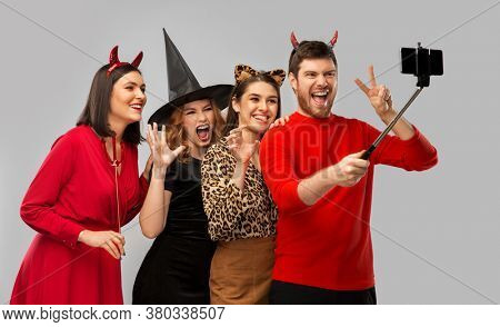 friendship, holiday and people concept - group of happy smiling friends in halloween costumes of witch, devil and cheetah taking picture by smartphone on selfie stick over grey background
