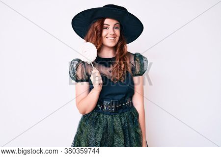 Young beautiful woman wearing witch halloween costume holding lollipop looking positive and happy standing and smiling with a confident smile showing teeth 