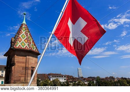 Swiss Flag On A Bridge In Basel (switzerland), Tower And City Skyline Against Bright Blue Sky