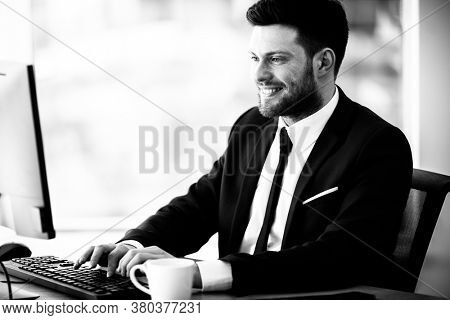 Business succsess concept. Happy smiling young businessman sitting in office and working on computer. Man in suit indoors on glass window background. Black and white, monochrome