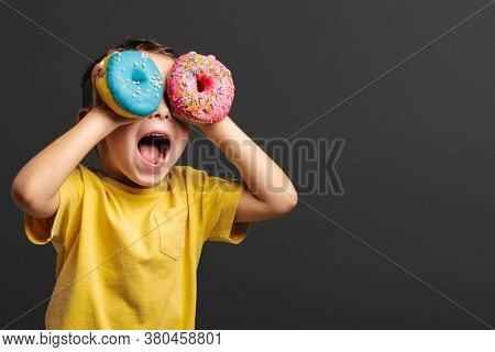 Happy cute boy is having fun played with donuts on black background wall. Bright photo of a boy. Colored donuts