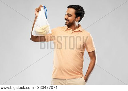 food, sustainability and eco living concept - happy smiling young indian man in polo t-shirt holding reusable string bag with bananas over grey background