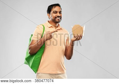 sustainability, consumerism and eco friendly concept - happy smiling indian man with green reusable canvas shopping bag and disposable box with takeaway food on grey background