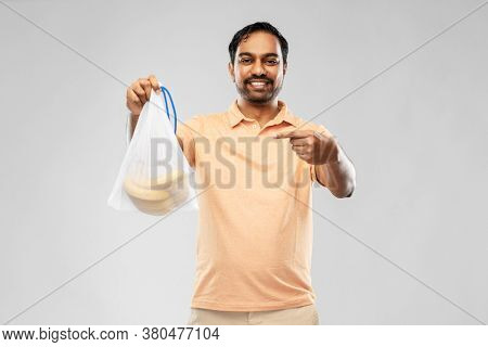 food, sustainability and eco living concept - happy smiling young indian man in polo t-shirt showing reusable string bag with bananas over grey background