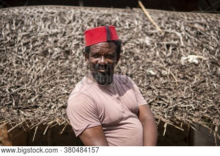 An African Older Man In Red Muslim Taqiyyah Fez Hat Posing With A Stick For Lame People On Yard Near