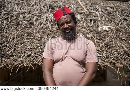 An African Older Man In Red Muslim Taqiyyah Fez Hat Posing With A Stick For Lame People On Yard Near