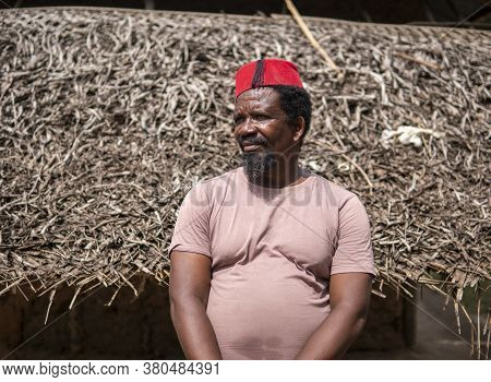 An African Older Man In Red Muslim Taqiyyah Fez Hat Posing With A Stick For Lame People On Yard Near