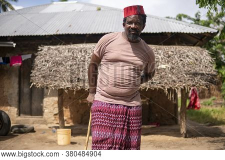 An African Older Man In Red Muslim Taqiyyah Fez Hat Posing With A Stick For Lame People On Yard Near