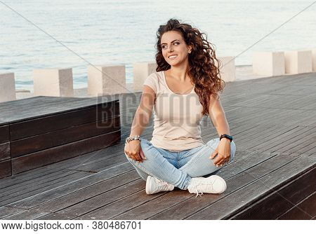 Young Women Wearing T-shirt And Jeans Sits On The Wooden  Bench And Smile