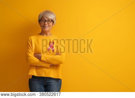 Senior woman on color yellow background. Pink ribbon like a symbol of breast cancer awareness. Support people living with tumor illness.
