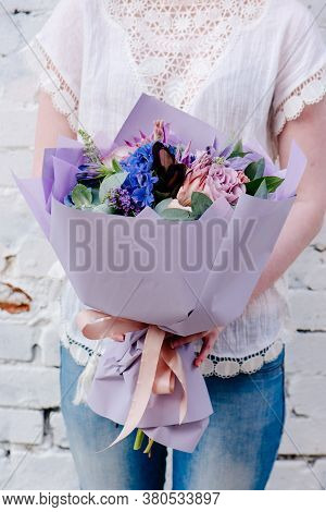A Bouquet Of Flowers, Packed In Purple Paper In The Hands Of A Woman On A Light Background. Bouquet 