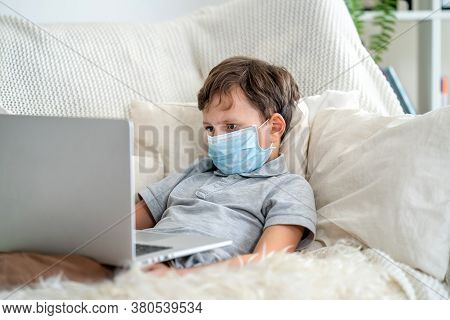 Focused Boy In A Mask, Lying On The Couch With A Laptop During The Quarantine Period. He Does Not Go