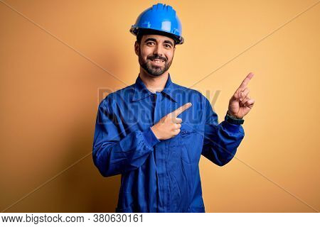 Mechanic man with beard wearing blue uniform and safety helmet over yellow background smiling and looking at the camera pointing with two hands and fingers to the side.