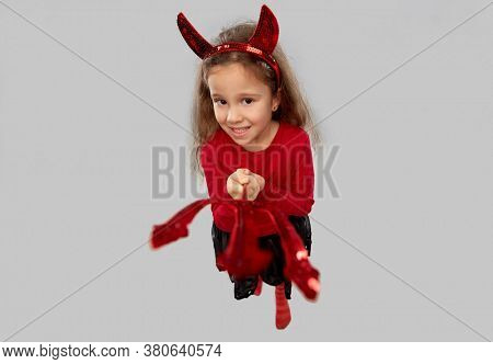 halloween, holiday and childhood concept - smiling girl in party costume with red devil's horns poking trident to camera over grey background