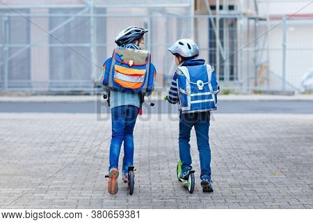 Two School Kid Boys In Safety Helmet Riding With Scooter In The City With Backpack On Sunny Day. Hap