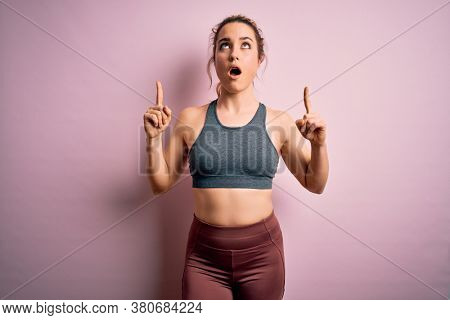 Young beautiful blonde sportswoman doing sport wearing sportswear over pink background amazed and surprised looking up and pointing with fingers and raised arms.