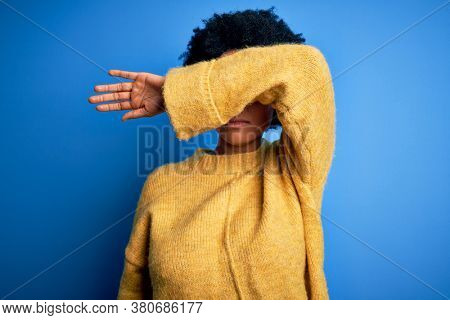 Young beautiful African American afro woman with curly hair wearing yellow casual sweater covering eyes with arm, looking serious and sad. Sightless, hiding and rejection concept