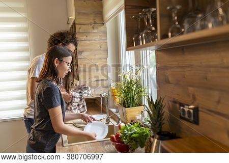 Young Couple In Love Doing The Washing Up And Drying Together After Lunch; Woman Washing While Man I
