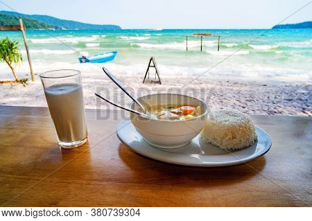 Thai Cuisine. A Plate Of Soup With Coconut Milk And Boiled Rice On The Table Against The Beautiful S