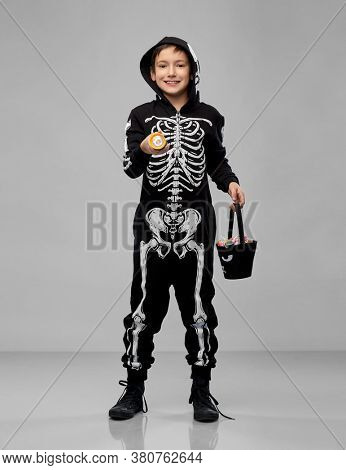 halloween, holiday and childhood concept - boy in black costume of skeleton with bucket of candies and flashlight trick-or-treating over grey background