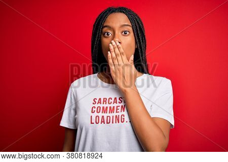 Young african american woman wearing sarcasm coments text on t-shirt over red background cover mouth with hand shocked with shame for mistake, expression of fear, scared in silence, secret concept