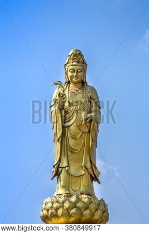 Golden Statue Of The Goddess Of Mercy  Guanyin Or Guan Yin Standing On The Lotus On Blue Sky Backgro