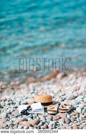 Beach Hat On Opened Book With Sunscreen And Shoes On Pebble Beach