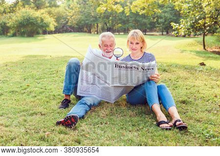 Happy Couple Elder Reading The Business Newspaper Together Through A Magnifying Glass. Elderly Picni