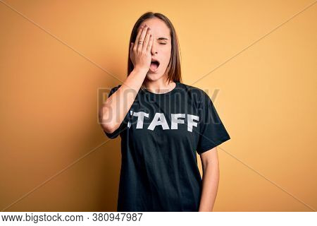 Young beautiful worker woman wearing staff uniform t-shirt over isolated yellow background Yawning tired covering half face, eye and mouth with hand. Face hurts in pain.