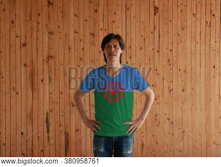Man Wearing Romani People Flag Color Shirt And Standing With Akimbo On The Wooden Wall Background. B