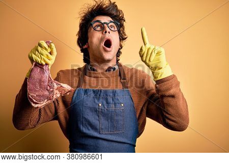 Young handsome butcher man holding meet steak standing over isolated yellow background amazed and surprised looking up and pointing with fingers and raised arms.