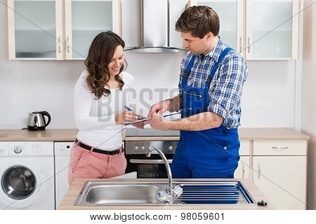 Woman Writing On Clipboard With Plumber In Kitchen Room