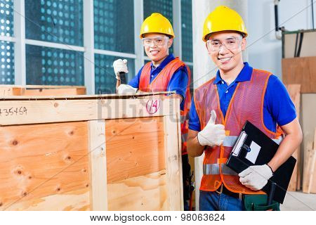 Two Asian Indonesian industrial or construction workers controlling with a checklist a delivery on a tower building site and open a wood box or cargo container with a crowbar 