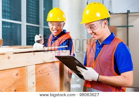 Two Asian Indonesian industrial or construction workers controlling with a checklist a delivery on a tower building site and open a wood box or cargo container with a crowbar 