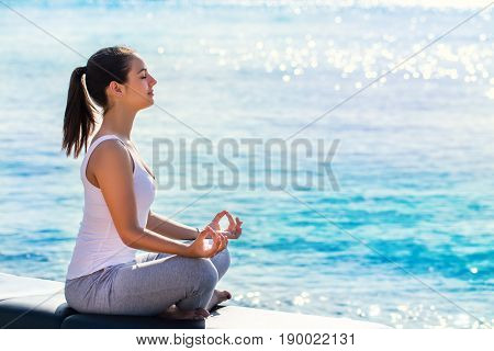 Full length portrait of young woman meditating next to sea. Side view of girl doing yoga exercise in early morning sun with bright reflection on water surface.