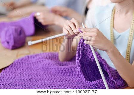 Fine workmanship. The focus being on dainty female hands knitting a scarf with purple threads in a chunky knit manner.