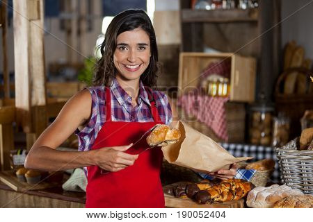 Portrait of smiling female staff packing sweet food in paper bag at counter in bakery shop