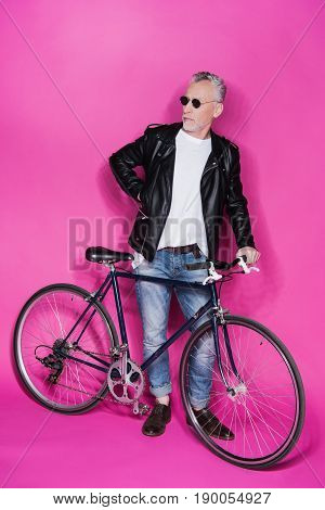 Fashionable Senior Man Wearing Leather Jacket And Sunglesses Standing With Bicycle Isolated On Pink